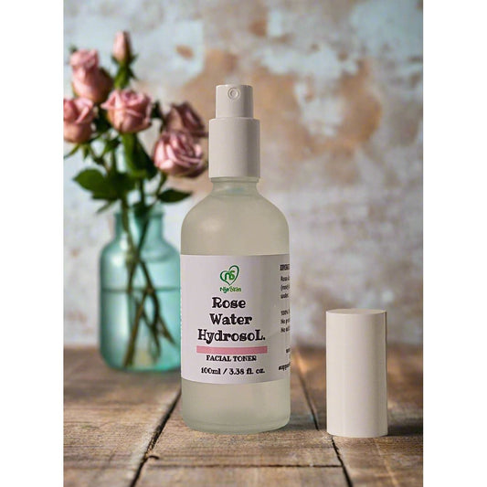 A clear spray bottle labeled 'Rose Water Hydrosol' with a capacity of 100ml, displayed on a wooden surface with a vase of pink roses in the background.